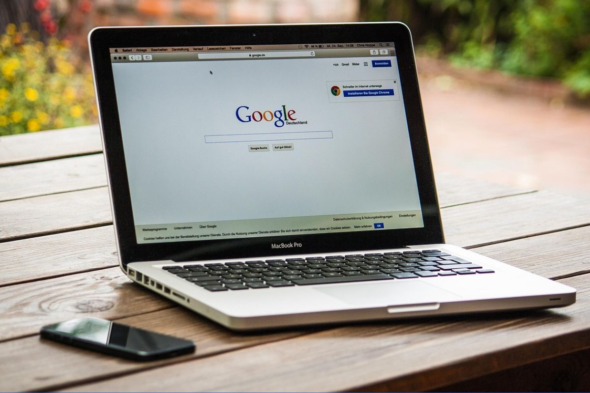 Notebook MacBook Pro exibindo a página inicial do Google, sobre uma mesa de madeira ao ar livre.