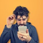 Homem tentando enxergar o celular com dificuldade, representando dúvidas sobre qual a melhor operadora de celular.